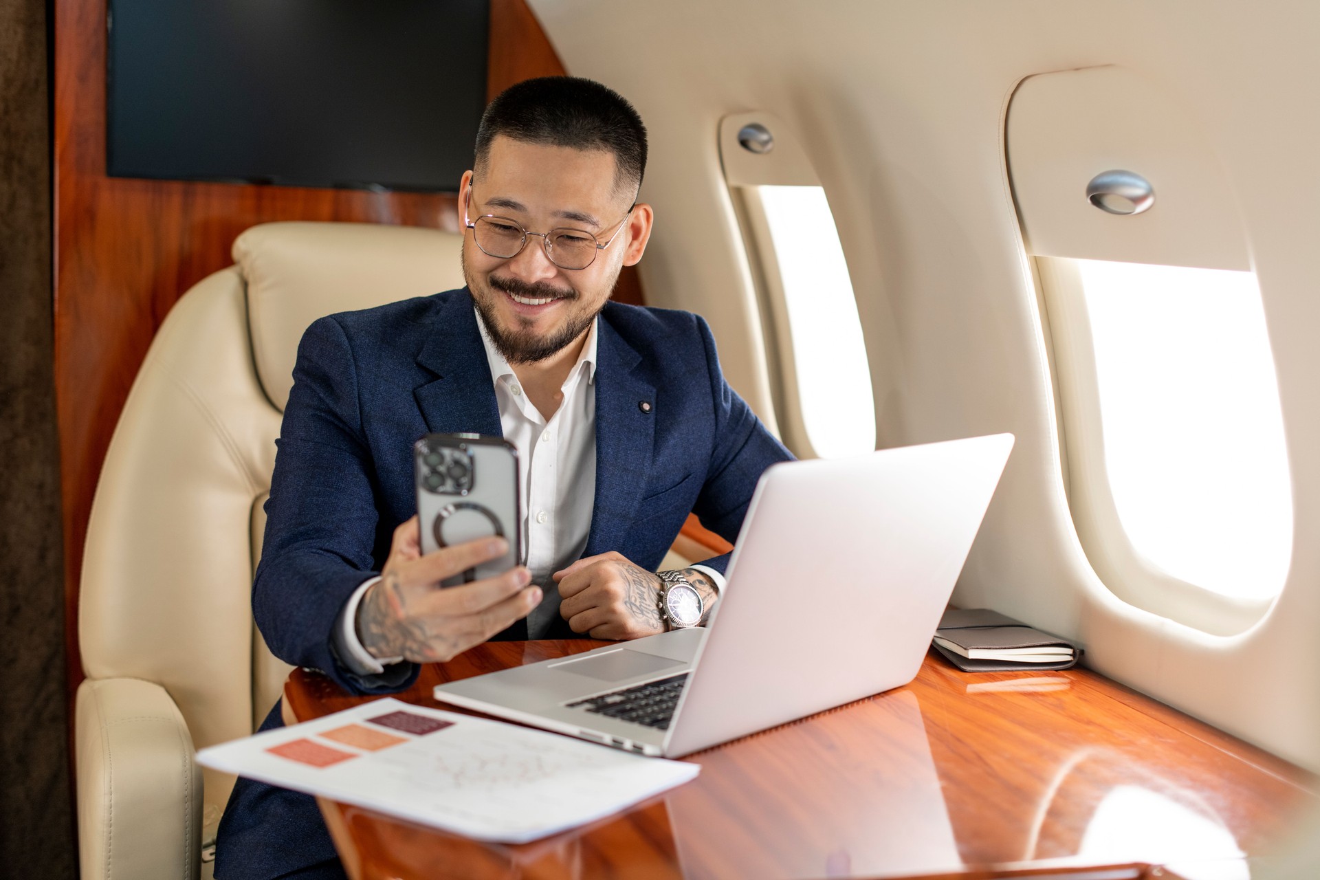 successful young asian businessman is sitting in private luxury jet with laptop and chatting via smartphone video call
